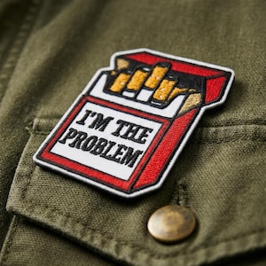 May include: Embroidered patch featuring a cigarette pack design with the text "I'M THE PROBLEM" in black. The patch has a red border and is set against an olive green fabric background. A brass button is visible below the patch.