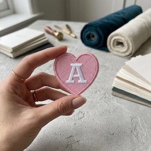 May include: A heart-shaped embroidered patch in light pink featuring a white capital letter A in the center. The patch has a stitched border and a textured fabric surface, showcasing a clear and bold design suitable for various textile applications.