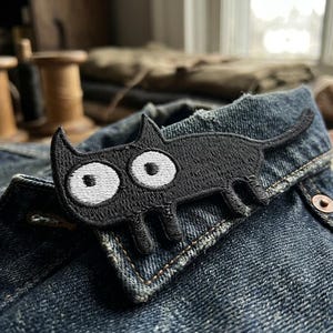 May include: A black embroidered cat patch with large white eyes is affixed to a denim jacket. The patch is in a cartoon style, with a simple outline and a small tail. The jacket is a medium wash denim with visible stitching and a button closure.