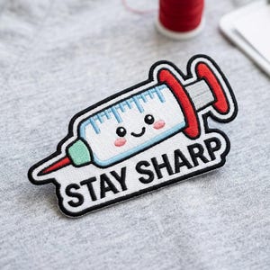 May include: Embroidered patch featuring a smiling syringe with blue and red accents, and the text "STAY SHARP" in black. The patch has a white background with a black border. The patch is on a gray surface.