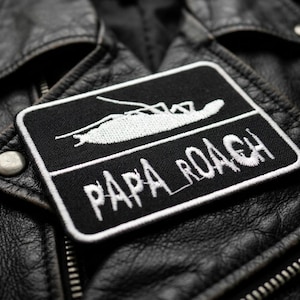 May include: A black and white embroidered patch featuring the text "PAPA ROACH" and a stylized insect design. The patch is rectangular with a white border and is affixed to a black leather jacket. The jacket has silver metal snaps and a zipper.