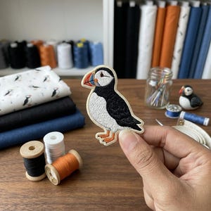 May include: An embroidered iron-on patch featuring a puffin with a black and white body, orange feet, and a multi-colored beak with red, blue, and yellow details. The patch has a cream-colored border and is held against a background of sewing supplies.