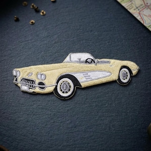 May include: An embroidered patch in the shape of a vintage convertible sports car. The car is light yellow with white side panels, silver-grey detailing on the grille and bumpers, and black and white wheels. The interior features a black steering wheel.