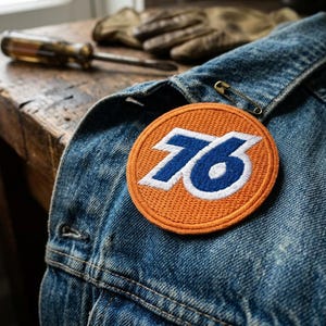 May include: A denim jacket with a circular orange patch featuring the number "76" in white and blue. A screwdriver and work gloves rest on a wooden surface in the background. A safety pin secures the jacket.