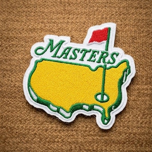 May include: Embroidered patch featuring the Masters Tournament logo. The patch is shaped like a map of the United States, with a yellow interior and green border. The word "MASTERS" is embroidered in green, and a red flag is visible.