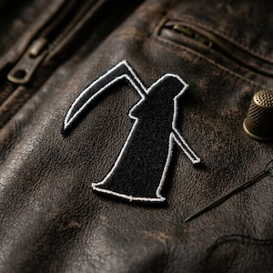 May include: A black Grim Reaper patch with white trim, sewn onto a distressed brown leather jacket. A gold thimble and needle with black thread are also visible, suggesting a sewing or crafting theme.