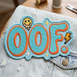 May include: Embroidered patch with the word "OOF" in turquoise, outlined in orange. The patch features a melting smiley face, stars, and a lightning bolt. The patch is on a denim jacket.