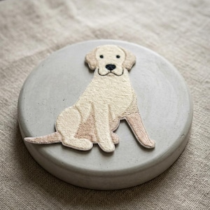 May include: A light beige embroidered dog patch is centered on a round, light gray concrete coaster. The dog is a Golden Retriever, with a black nose and smiling mouth. The coaster is on a tan linen surface.