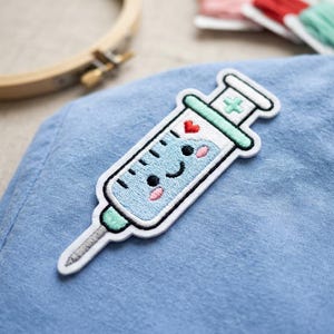 May include: Embroidered patch of a smiling syringe with a heart and cross design. The syringe is light blue with a white outline and green accents. The patch is on a blue fabric background.