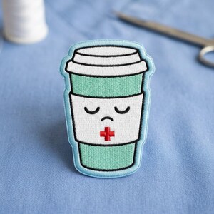 May include: Embroidered patch featuring a teal coffee cup with a sad face and a red cross. The patch has a white paper sleeve and a light blue border. The background is a light blue fabric, with a spool of white thread and scissors.