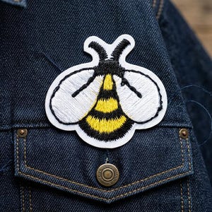 May include: Embroidered bee patch with white wings, black and yellow striped body, and black antennae. The patch is sewn onto a dark blue denim jacket pocket. The jacket has a button closure and visible stitching.