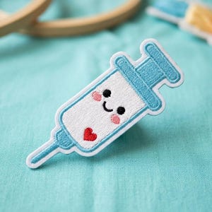 May include: An embroidered patch in the shape of a syringe with a smiling face, black eyes, and a red heart. The syringe is white and light blue, with a white border. The background is a light blue fabric.