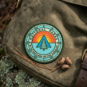 May include: A round National Parks patch with a compass design, featuring a tree and a sunset over a blue lake. The patch is on a green backpack. Text includes "NATIONAL PARKS" and "IN THE DISCOVERY WE FIND OURSELVES".