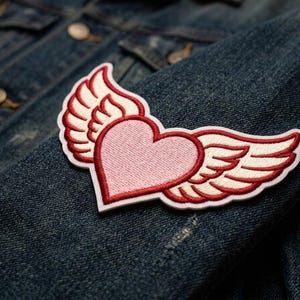 May include: Embroidered patch featuring a pink heart with white and red wings. The patch is sewn onto a blue denim jacket. The heart and wings design is a popular symbol of love and freedom, often used in fashion and crafts.