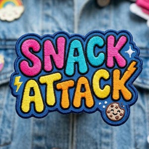 May include: Embroidered patch with the words "SNACK ATTACK" in pink, teal, yellow, and orange, outlined in blue. A cookie graphic is at the bottom right. The patch is on a denim jacket.