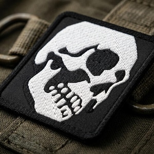 May include: A square black patch with a white skull design. The skull has detailed features, including eye sockets, teeth, and a defined jawline. The patch is embroidered and has a black border. The background is a textured olive green fabric.