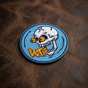 May include: Round embroidered patch featuring a white skull with yellow eyes and the letters "WTF" in yellow. The skull is set against a light blue background with concentric lines. The patch is on a brown leather-like surface.