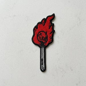 May include: Embroidered patch featuring a stylized red flame with a black outline. The flame design includes a skull and the text "OL' SKULL". The patch has a black stem and is likely for clothing or accessories.