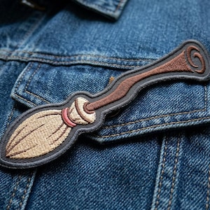 May include: Embroidered patch in the shape of a broom, featuring tan bristles, a pink band, and a brown handle with a swirl design. The patch is outlined in gray and is placed on a denim jacket.