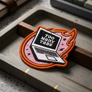 May include: Embroidered patch featuring a laptop with the text "Too Many Tabs" on a black screen. The patch has a pink and orange flame design around the laptop. The patch is on a gray tray with a wooden accent.