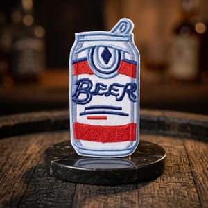 May include: Embroidered patch in the shape of a beer can. The design features a blue and red color scheme with the word "BEER" in a cursive font. The patch is on a dark marble surface.