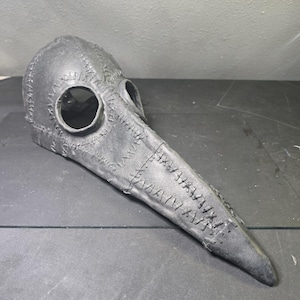May include: A gray plague doctor mask with a long, curved beak and large eye holes. The mask is made of a textured material and features visible stitching along the seams, giving it a handcrafted appearance. The mask is designed to cover the entire face.