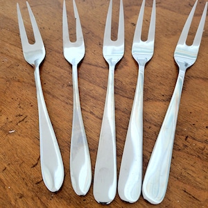 May include: Five silver serving forks with a two-tine design are arranged on a wooden surface. The forks have a polished, reflective finish, and the tines are evenly spaced. The handles have a teardrop shape.