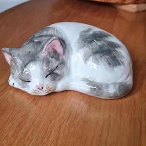 May include: A ceramic ornament of a sleeping cat. The cat is white with grey markings and pink ears. It is lying on a wooden surface. A wicker basket is in the background.