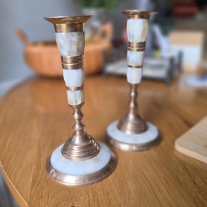 May include: A pair of brass candlesticks featuring inlaid white mother-of-pearl segments along the stems and circular bases. The metal has a warm, polished finish, and the white shell pieces are arranged in a grid pattern on the candle holders.