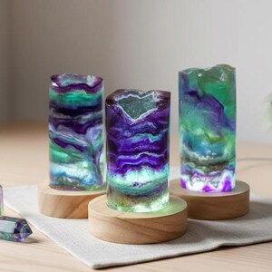 May include: Three illuminated, cylindrical resin lamps with a layered, colorful design of purple, green, and white. Each lamp sits on a round wooden base. Several small, faceted crystal stones are in front of the lamps.