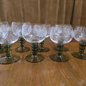 May include: A set of seven clear glass goblets with etched floral patterns on the bowls. Each goblet features a distinctive dark olive green stem with a stacked ball design and a matching circular base, creating a cohesive glassware collection.