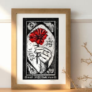 Op de afbeelding: Ingelijste kunstdruk met een rode bloem die door een hand wordt vastgehouden, met de tekst "It's an Old Song" en "And we're gonna sing it again." Het kunstwerk is in zwart-wit met een houten lijst.