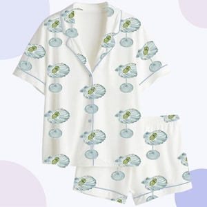 Könnte beinhalten: Weißes Pyjama-Set mit Muschelmuster. Das kurzärmelige Oberteil und die Shorts zeigen ein sich wiederholendes Design aus blauen und grünen Muscheln auf weißem Hintergrund, mit hellblauen Paspeln.