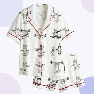 Könnte beinhalten: Weißes Pyjama-Set mit einer sich wiederholenden Schwarz-Weiß-Illustration von Katzen, die mit Gewichten, Boxsäcken und Springseilen trainieren. Das Oberteil und die Shorts haben rote Paspeln.