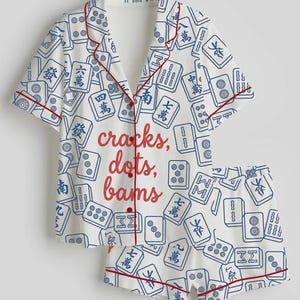 May include: White pajama set with a blue Mahjong tile pattern and red trim. The short-sleeved top and shorts feature the text "cracks, dots, bams" in red. The set is made of a soft, lightweight fabric.