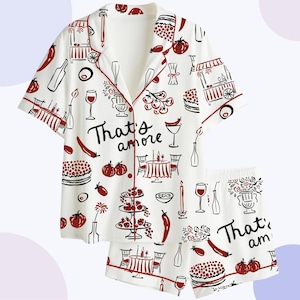 May include: White pajama set with a red trim and a repeating pattern of Italian food illustrations. The set includes a short-sleeved button-down shirt and matching shorts. The phrase "That's amore" is printed on the shirt and shorts.