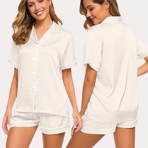 May include: Cream-colored pajama set featuring a short-sleeved button-down shirt and matching shorts. The set has white piping along the edges of the collar, sleeves, and shorts. The fabric appears to be a smooth, satin-like material.