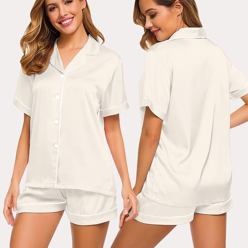 May include: Cream-colored pajama set featuring a short-sleeved button-down shirt and matching shorts. The set has white piping along the edges of the collar, sleeves, and shorts. The fabric appears to be a smooth, satin-like material.