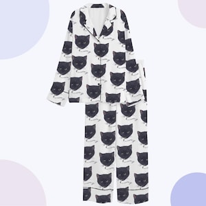 May include: White pajama set with a black cat face pattern. The long-sleeved top has a notched collar and button closure. The matching pants have a relaxed fit. Black trim accents the collar, cuffs, and pant hems.