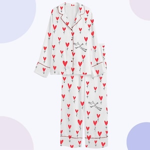 Puede incluir: Conjunto de pijama blanco con estampado de corazones rojos. La parte superior de manga larga tiene una botonadura y ribetes rojos. Los pantalones a juego también presentan el diseño de corazón. Una ilustración caprichosa de una figura está presente.