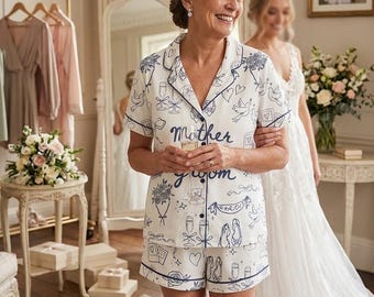 May include: A white pajama set with a blue outline design and the words "Mother of the Groom". The set includes a button-down shirt and shorts. The design features illustrations of doves, champagne flutes, and shoes. The woman is smiling.