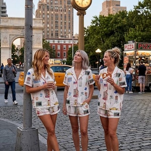 Könnte beinhalten: Drei Personen tragen passende weiße Pyjama-Sets mit einem New York City-Motivdruck und posieren auf einer Stadtstraße. Die Pyjamas haben rote Verzierungen und zeigen Illustrationen von Gebäuden, Taxis und Brezeln.