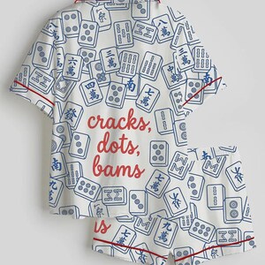 May include: White pajama set with a pattern of blue Mahjong tiles. The set includes a short-sleeved top and shorts, both trimmed with red piping. The words "cracks, dots, bams" are printed in red on the top.