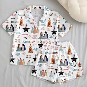 May include: White pajama set featuring a repeating pattern of illustrations and text from the musical "Hamilton." The set includes a short-sleeved button-up shirt and matching shorts. The design incorporates various characters, quotes, and symbols from the show.