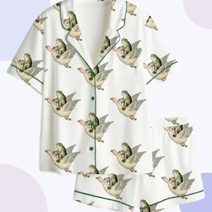 May include: White pajama set with a whimsical pattern of green frogs riding geese. The short-sleeved top and shorts are trimmed with green piping. The set features a button-down shirt and elastic waist shorts.
