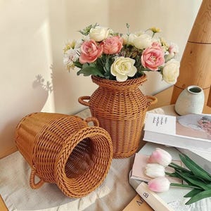 May include: Two brown woven wicker vases with small side handles. One vase stands upright and is filled with a bouquet of artificial pink and white roses, while the other vase is positioned on its side in front of the upright one.