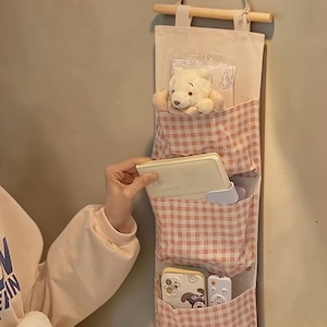 以下が含まれることがあります： ピンクと白のギンガムチェック柄のポケットが3つ付いた、オフホワイトの布製壁掛け収納オーガナイザー。木製の棒から吊り下げられており、小さなクマのぬいぐるみ、ノート、スマホケース、リモコンが収納されています。
