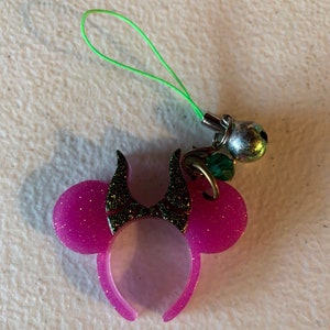 Puede incluir: Un dije de diadema de orejas de ratón rosa brillante con cuernos negros, un cordón verde y una campana plateada. El dije es un accesorio divertido para bolsos, llaveros u otros artículos.
