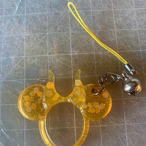Puede incluir: Un charm de resina amarilla con forma de orejas de ratón y detalles de purpurina. El charm está unido a un cordón amarillo con una campana y un cierre plateados. El charm está sobre una superficie gris con marcas numéricas.