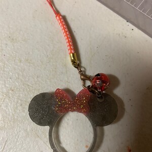 Puede incluir: Un dije de diadema de orejas de Minnie Mouse gris transparente con un lazo rojo brillante. El dije está unido a un cordón trenzado rojo y blanco con un cierre dorado y una pequeña cuenta roja y negra.